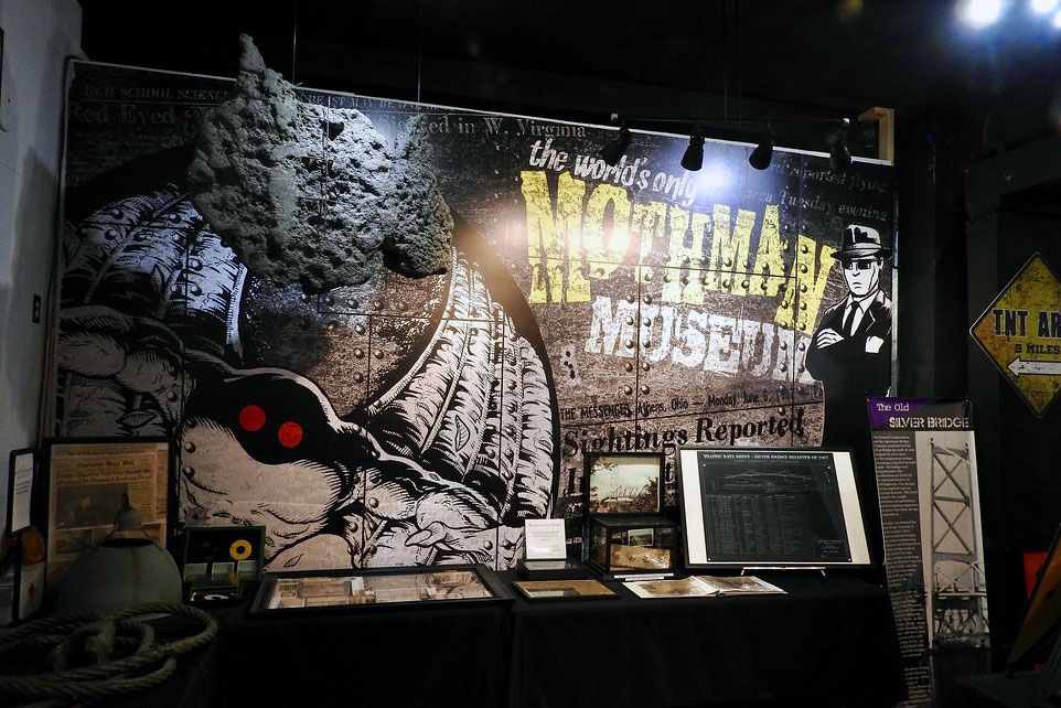 Display inside a museum with black walls and tables full of signs, photos, and a rope. The backdrop reads: “The world's only Mothman Museum” with cartoon illustrations of a Mothman with red eyes and a detective.