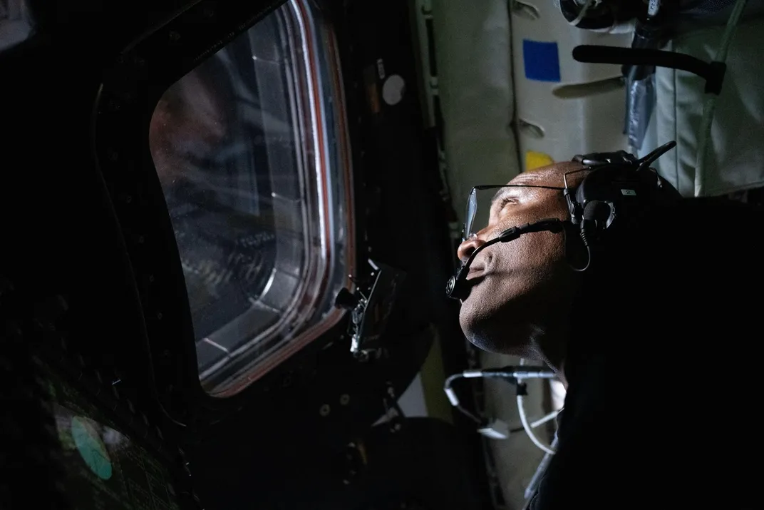 an astronaut wearing a headset looks out a spacecraft window in the dark