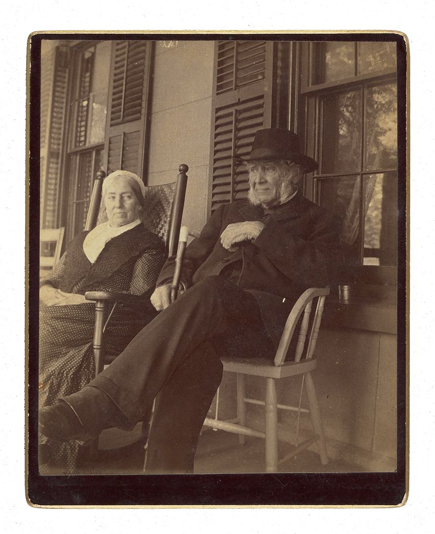 An older couple sitting on a porch. The man sits in a chair  in a suit and hat, holding a cane with his legs crossed. The woman has a bonnet on and sits in a rocking chair. 