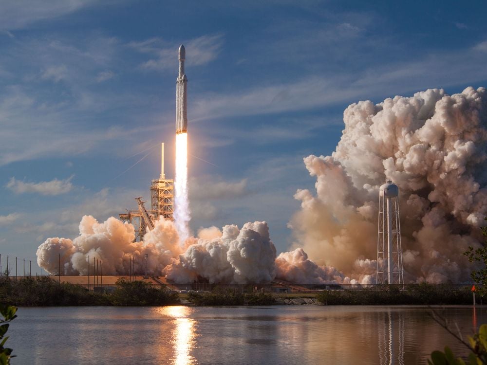 A rocket launches into a bright blue sky from a launchpad amid a great deal of smoke and near a body of water