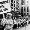 Woman holding a sign reading “Equal positions with equal pay” during an equal pay demonstration in a city plaza, with women seated behind her.