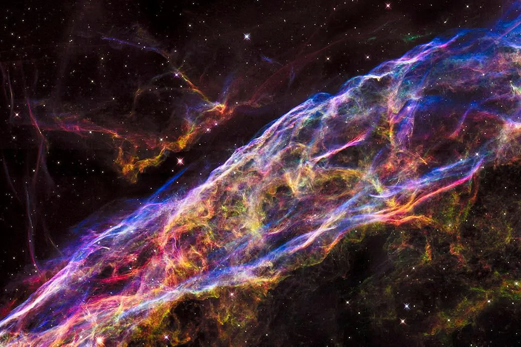 An image from 2015 of the Veil Nebula. The nebula is glowing with various intertwined shades of green, blue, red and yellow against a dark starry sky.  