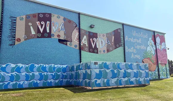Turquoise mural covering the entire side of a building, with a pattern of waves along the bottom, and a brown textile with the Spanish words Viva la Vida! in the center.