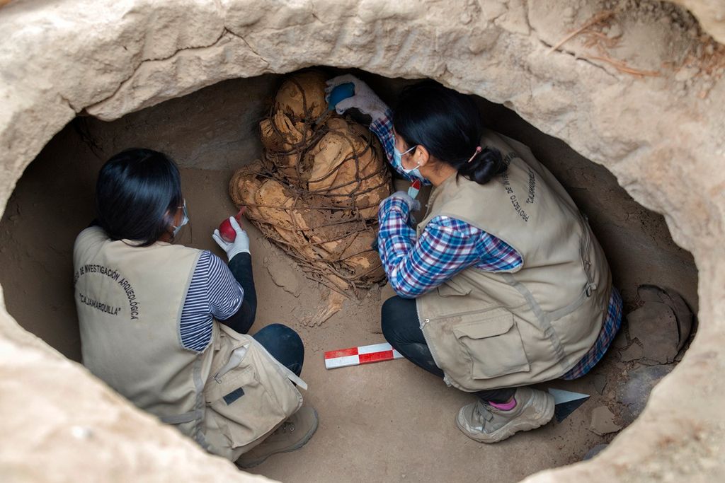 Two people kneel in an oval-sized opening and carefully brush the mummified body
