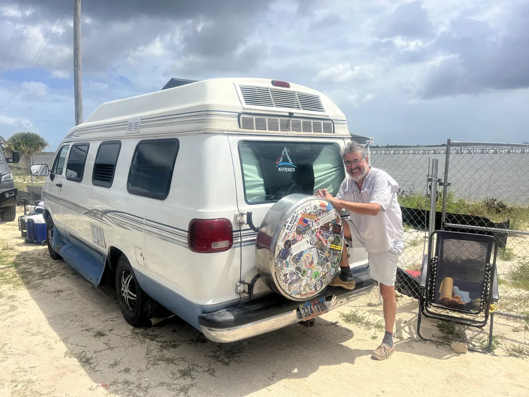 a man stands behind an old van with a spare tire on the back within a cover coated with stickers