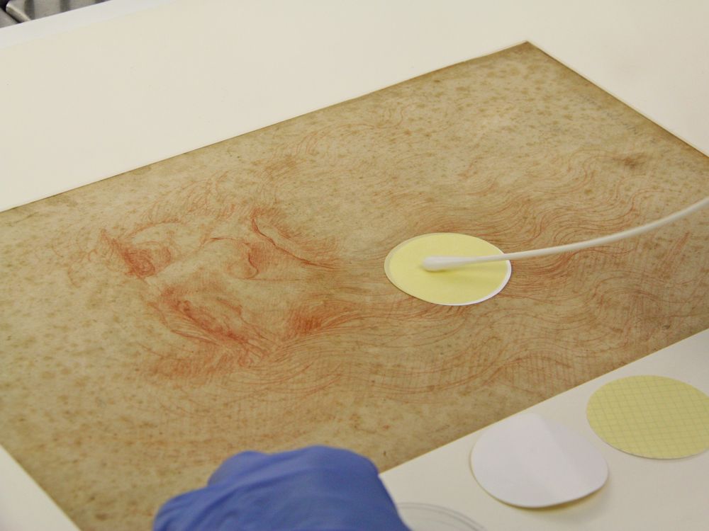 A pair of blue gloved hands gently wipe a bendy q-tip apparatus over a small circle of yellow paper that appears to be resting on the surface of the sketch, which is about the size of a normal sheet of paper