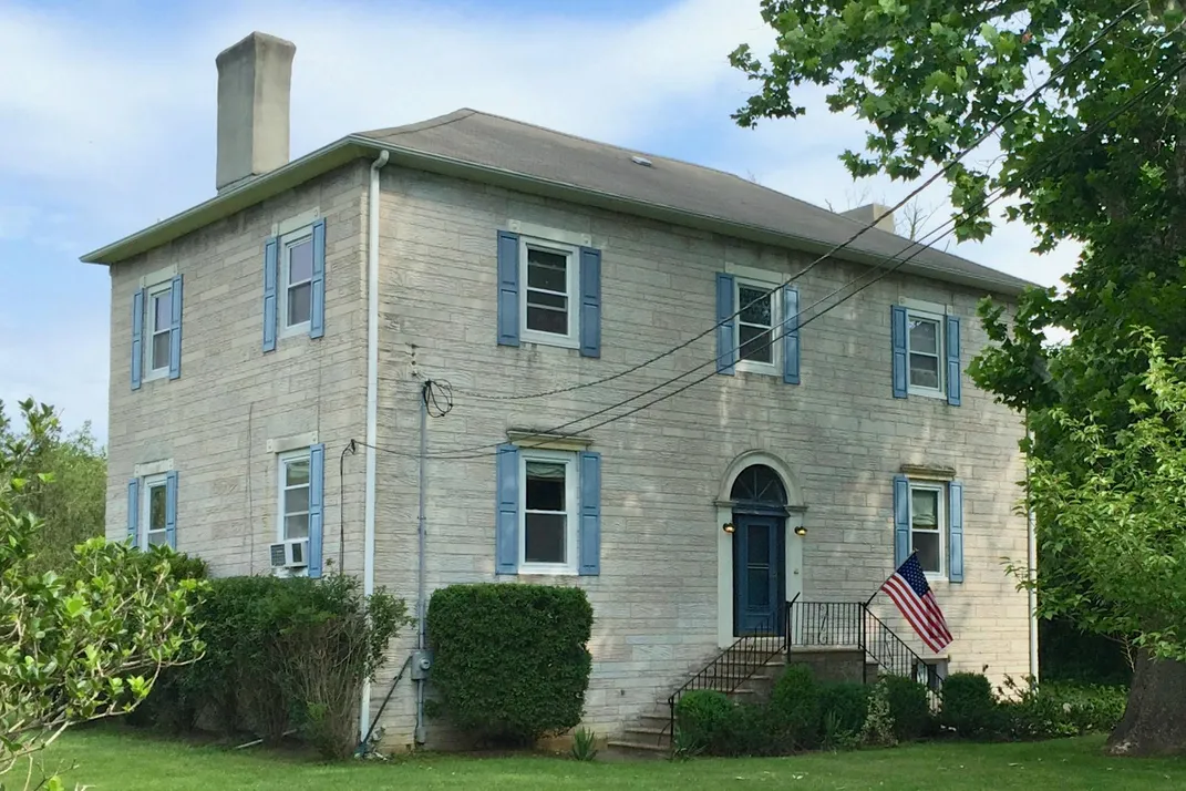 A white colonial home, spare with blue window shutters and surrounded by greenery, stands in front of a blue sky with an American flag hanging out front