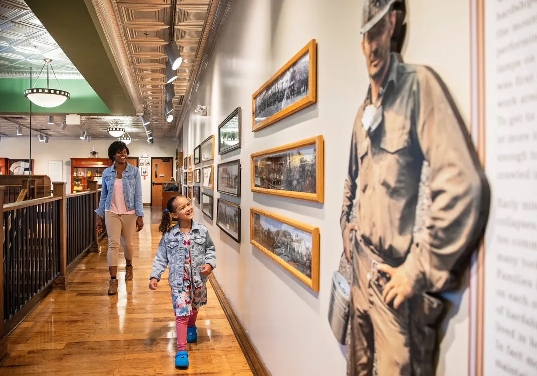 Interior museum gallery at Beckley Exhibition Coal Mine featuring historical photographs and mining exhibits along a hallway. Two visitors walk through the space, with a life-size image of a coal miner displayed prominently in the foreground.