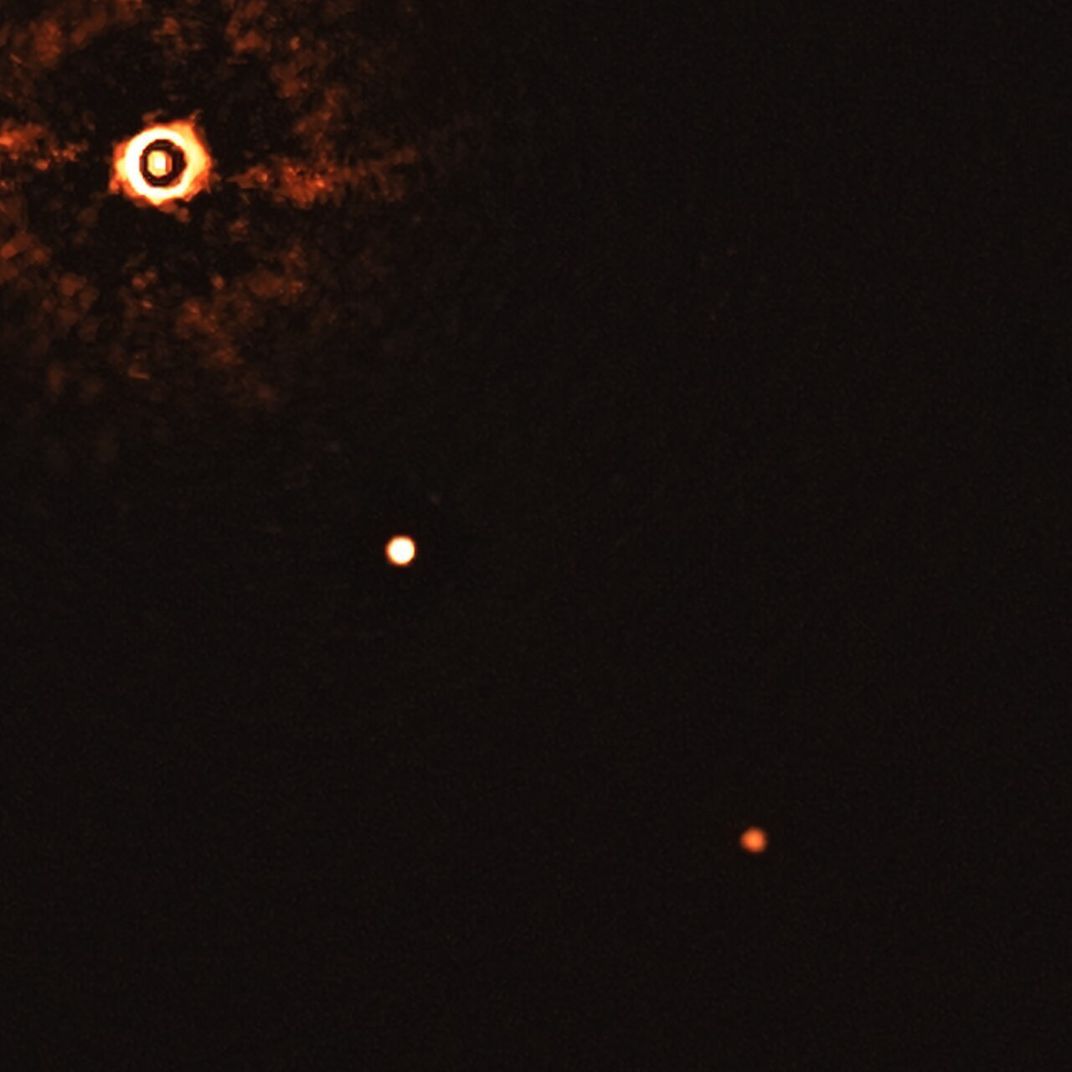 On the far left, a bright object that radiates light (the star); a bright white light near the center of the image is the larger exoplanet and a further, dimmer orange pinprick is the smaller planet