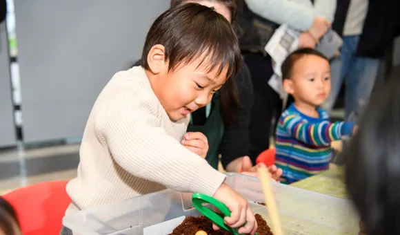 A child smiles as he leans over a plastic tub filled with dirt using a set of tweezers to move objects around in the dirt.
