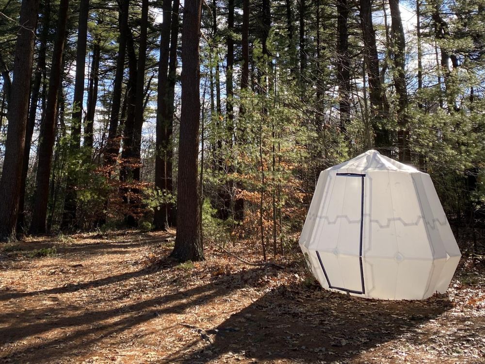 A white octogon-shaped origami tent sits in the woods in New England