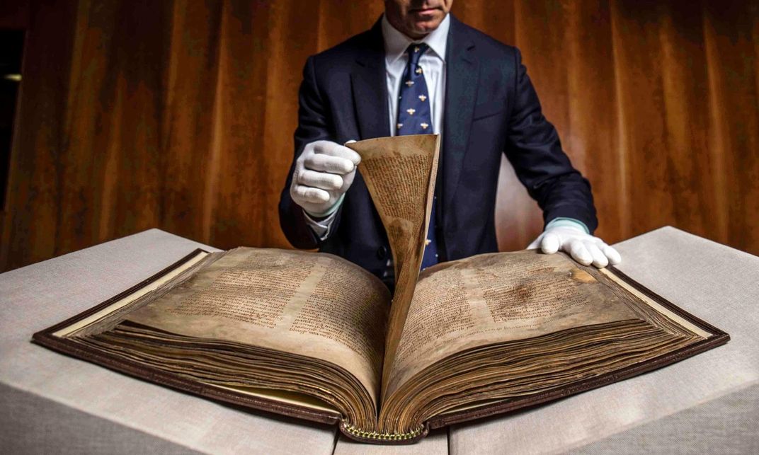 A person in a suit and tie wears white gloves and carefully turns one page of the book; the large book, with brown and yellowed vellum, is spread out open on an angled table