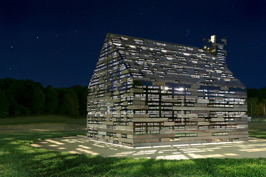 A cabin structure is illuminated from within, with light pouring out in between metal slats engraved with lines of poetry