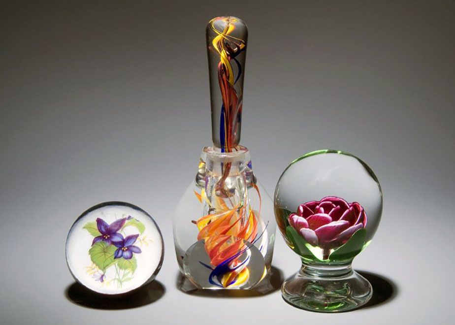 Three glass-blown pieces: an orb containing purple flowers with green leaves, a bottle with orange and purple streaks that look like flames, and an orb with a pink rose.