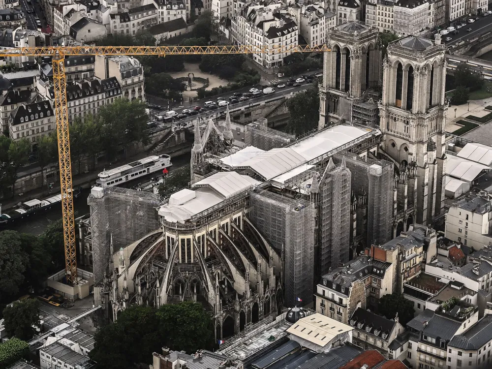 An aerial view of the exterior of the cathedral, its damaged roof mostly covered by white tarp. An enormous crane looms over the building