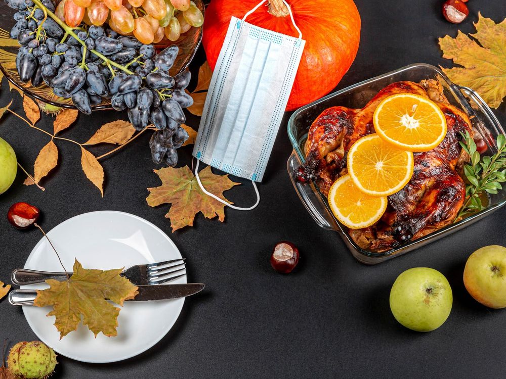 A photograph of a table taken from above. Table has dried leaves, a white plate with a fork and knife, a roasted chicken with orange slices in a glass tray. There is a a pumpkin with a blue disposable mask draped over the stem at the top of the image.