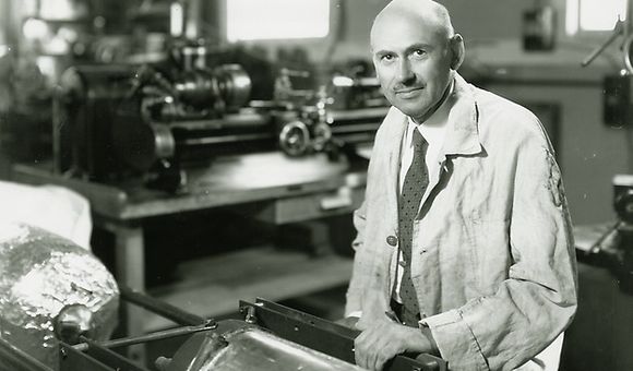 Black-and-white image of a white man in his early 50s. He is bald and has a mustache. Wearing a white lab coat, he is sitting at a work table supporting a large metal cylinder.