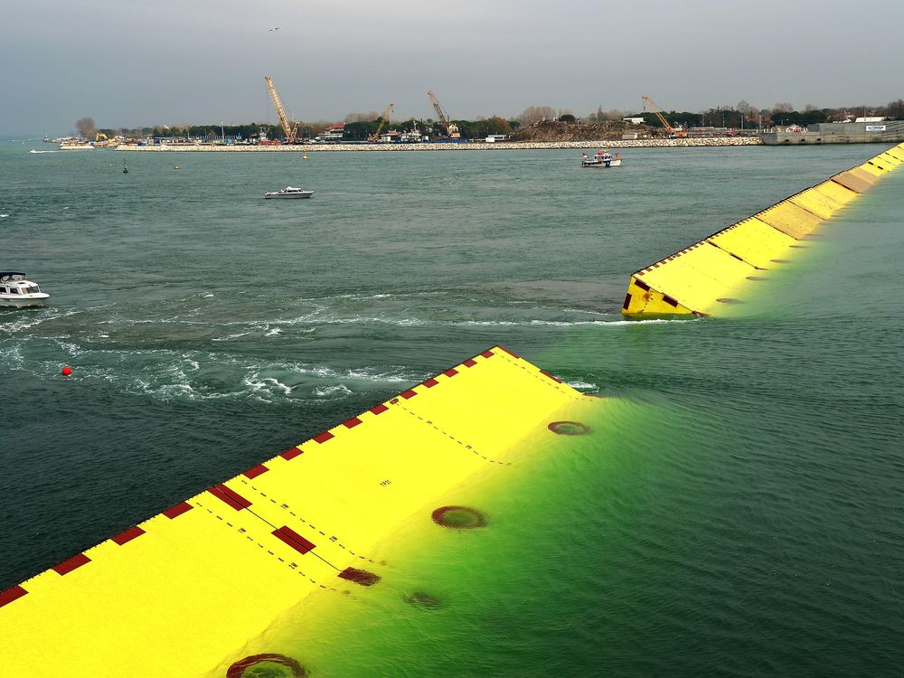 Near the shore, the flood barriers emerge from the water. They are large, bright yellow and triangularly shaped to deflect the water. There are several boats in the water nearby. Lido, a barrier island near Venice, is in the background. 