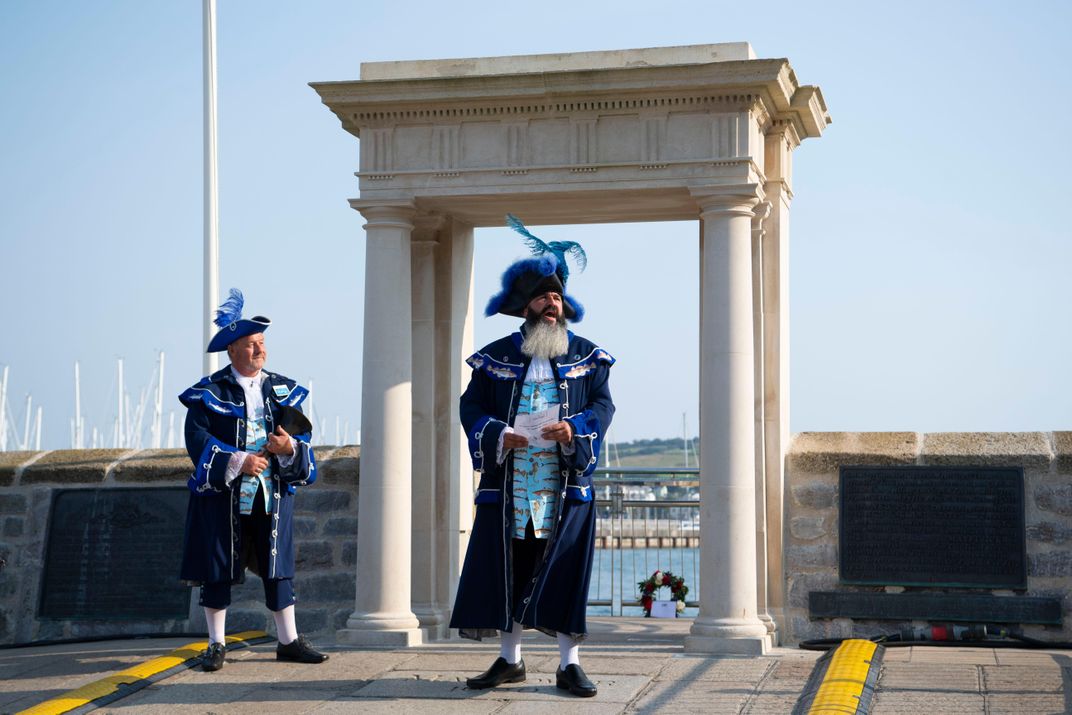 Two men in period costume, with blue feathers in their caps, speak in front of a neoclassical square archway that marks the spot where the Pilgrims set off for America, 400 years ago