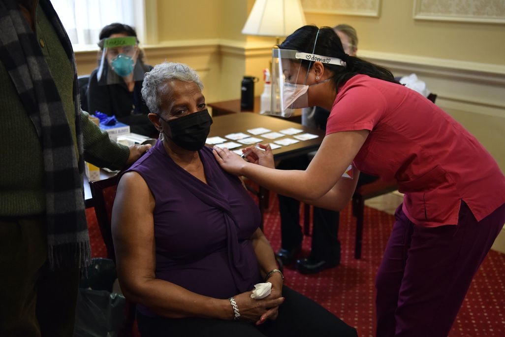 A woman sits in a chair and gets a vaccine shot from a woman wearing a face mask and shield