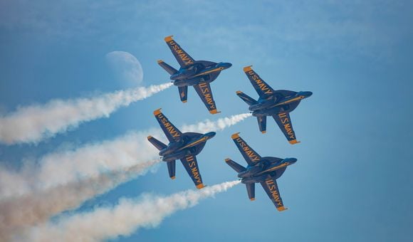 Four military jets fly in a diamond formation across a blue sky. On the underside of each jet's wings, "US Navy" is painted in bold yellow letters. White airshow smoke streams from each jet. The moon is visible in the sky.