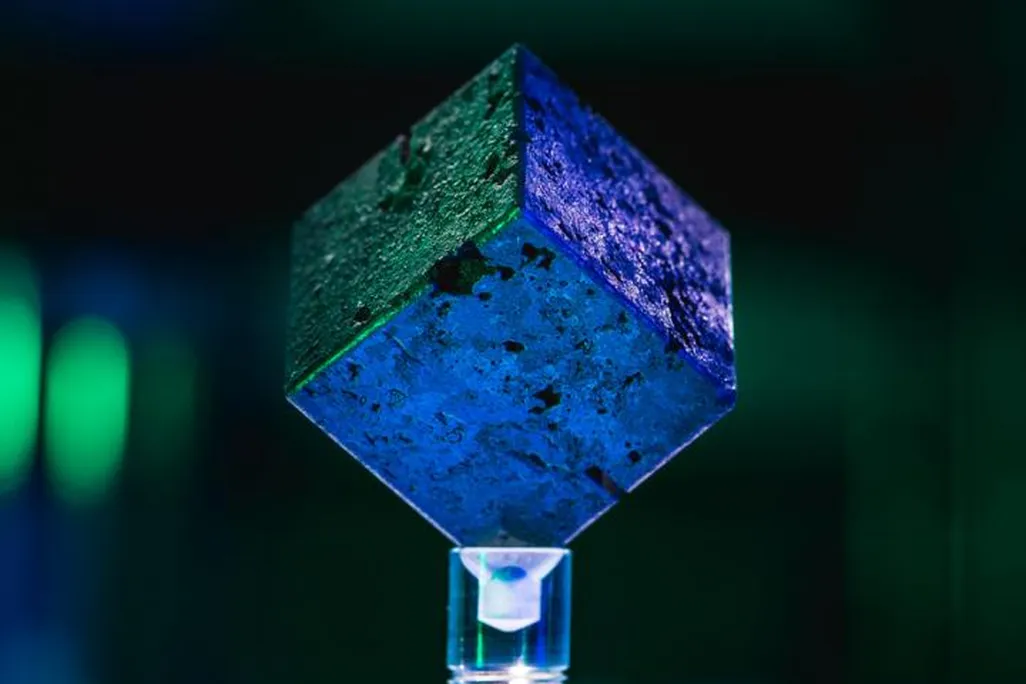A blue and green image of a small metal cube resting on a display, with colored lights in the background