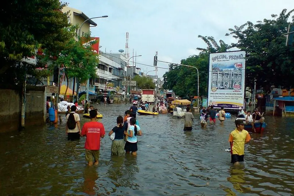 A view of a flooded marketplace in Jakarta. People are wading through knee-high water in the middle of a street. It looks like a typical, bustling street except that the street is entirely flooded.