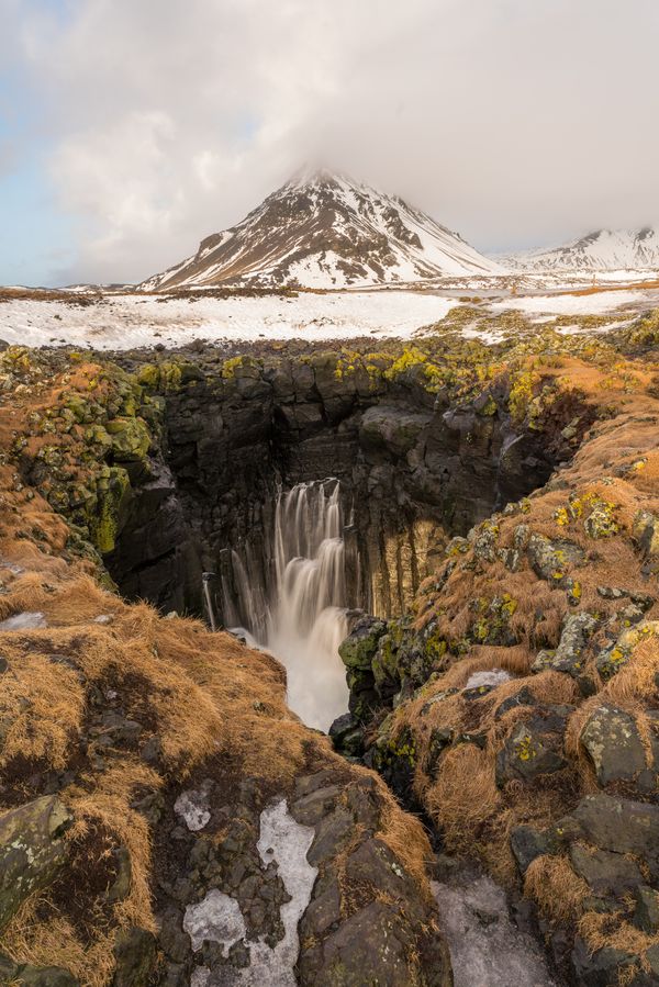 A coastal cliffside waterfall beneath a snow capped mountain in the Snaefellsnes Peninsula thumbnail