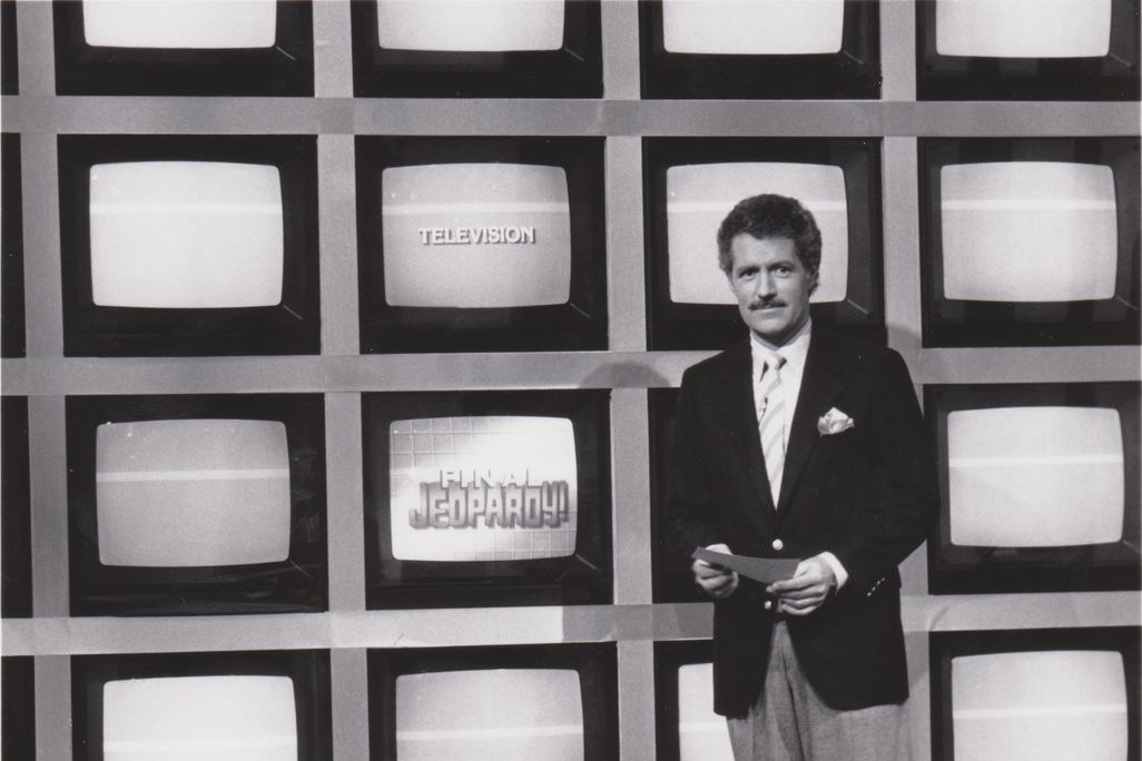A black and white image of a young Trebek, a white man in a suit and tie with dark hair and a mustache, standing in front of a grid of TV screens