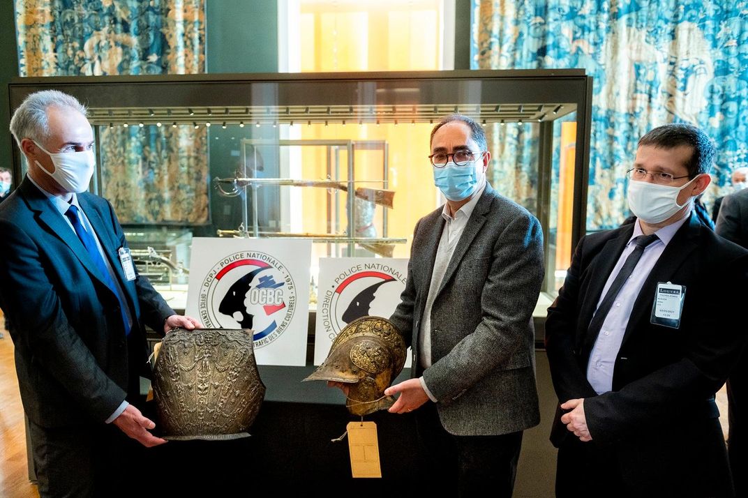 Three men stand around a table; two hold a piece of armor. The breastplate, left, is large and carved with a figure of a woman on its front; the helmet, right, is likewise intricate and has a curved brim that juts out