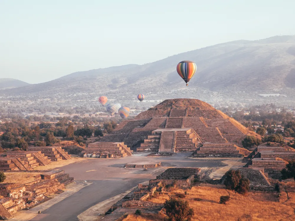 In an aerial view of an ancient city, a hot air balloon floats above an enormous stone temple with steps leading up to its peak