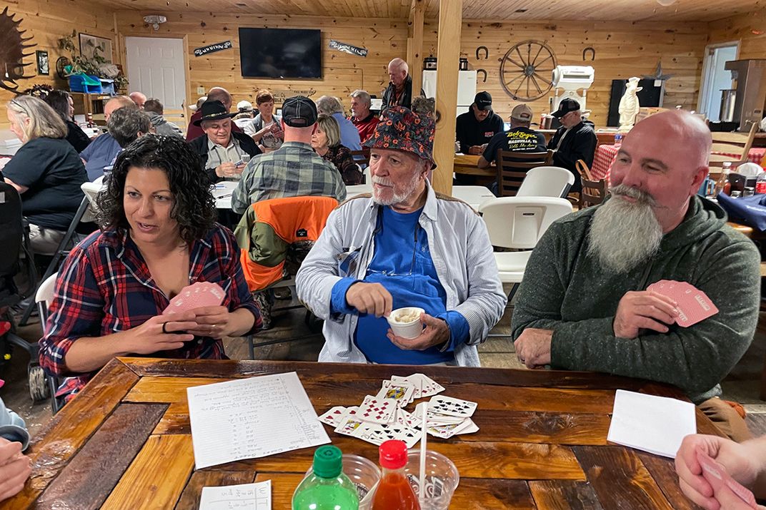 Three people sitting on one side of a table. Two are holding hands of playing cards; one is holding a cup of food.