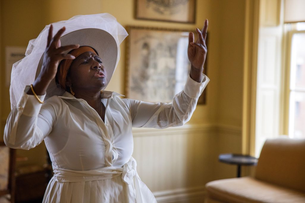 A black woman in a white dress and a large brimmed white hat stands in a room with yellow painted walls, raises her hands and closes her eyes, as though caught in the midst of dancing and singing