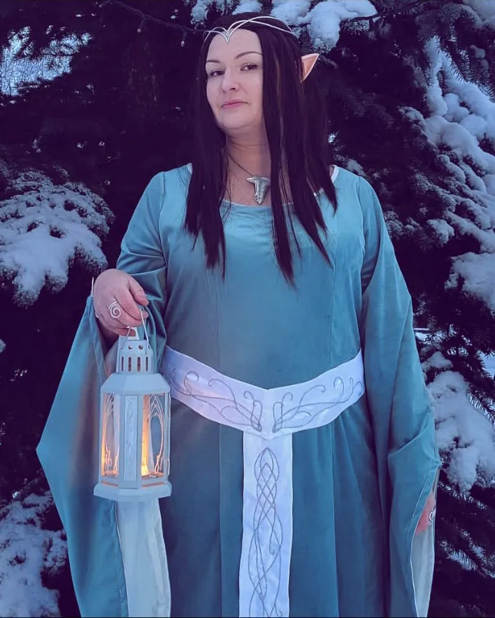 A woman wearing a flowing blue dress with white sash and thin silver crown poses in front of a snow-covered evergreen green, holding a lit lantern.