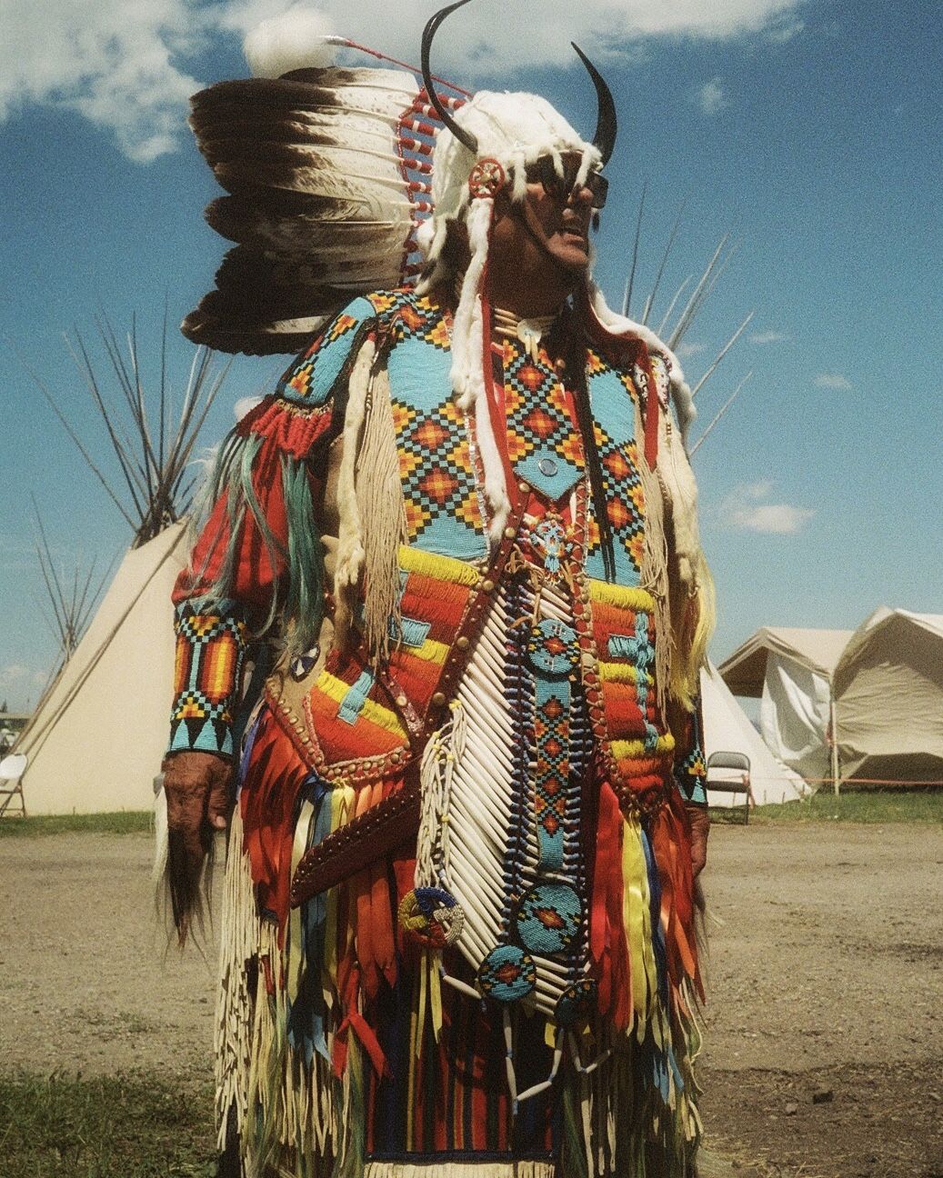a man stands in traditional attire, adorned with horns, feathers and colorful fabrics and ribbons.