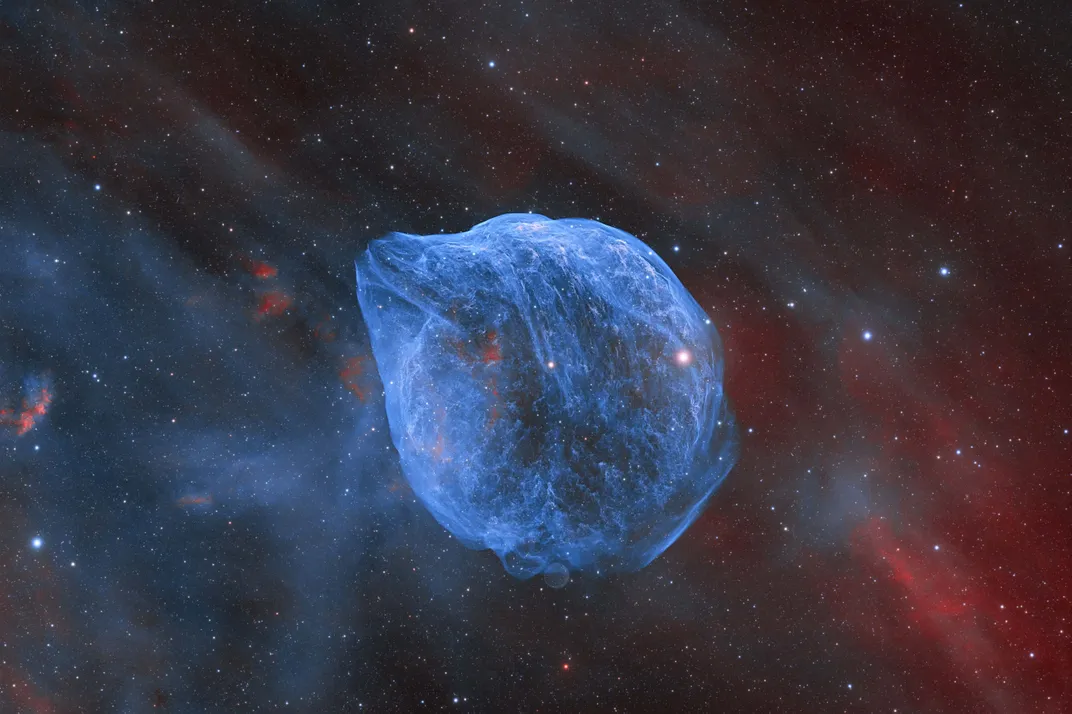 A blue nebula is looks like a dolphin's head with its nose pointing to the top left against a colored blue and red sky