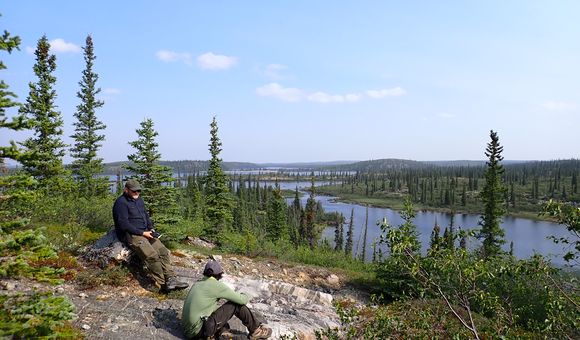 Two men sit on a rock bed on top of a forested hill over looking a remote lake.