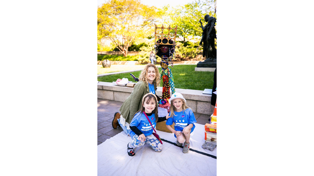 An adult and two children pose in front of a tall colorful figure wearing a golden crown. They are in a garden; greenery and a bronze sculpture can be seen in the background.