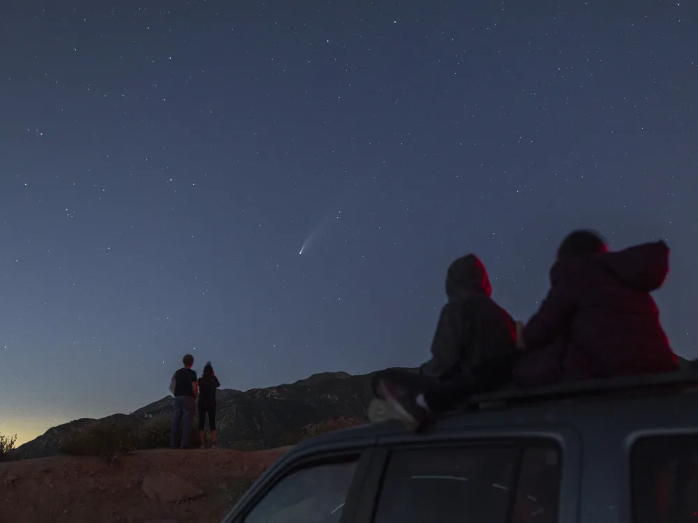 Outlines of figures, including two young people seated on top of a car, framed against twilight sky as they watch a comet streak across the sky