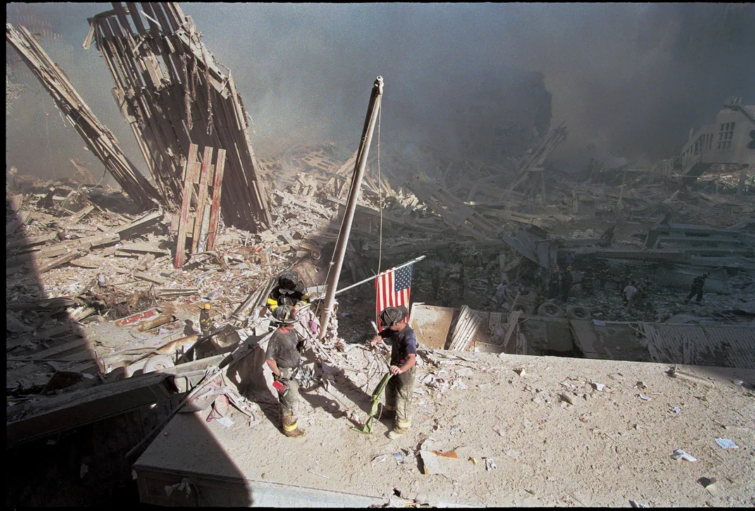 A horizontal sweeping view of the scene, with wreckage and smoke as far as the eye can see; center, the three small firemen work to remove the American flag from a pole and tie it to the large flagpole