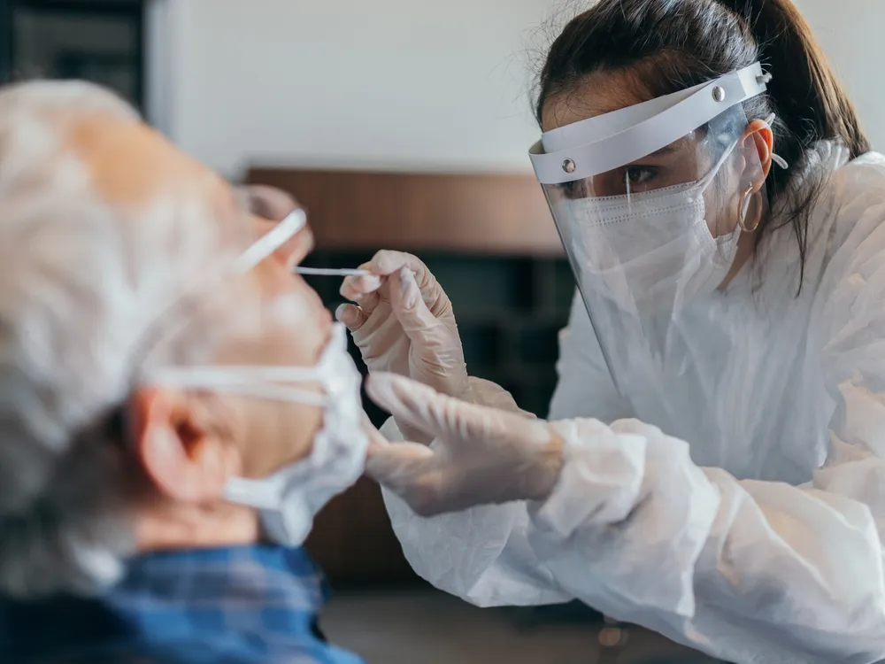 A female doctor in a mask and visor gives a nasal swab to a male patient.