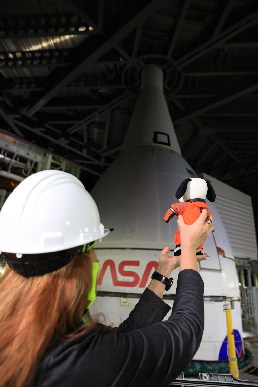 A woman wearing a hard-hat, holds a stuffed animal Snoopy facing the top of the SLS rocket.