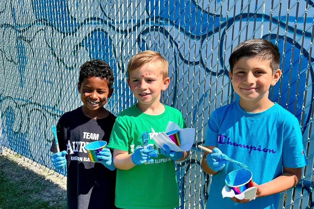 Three kids pose in front of painted blue waves of the mural, each holding a cup of blue paint and a paintbrush. Two of them have paint on their faces.