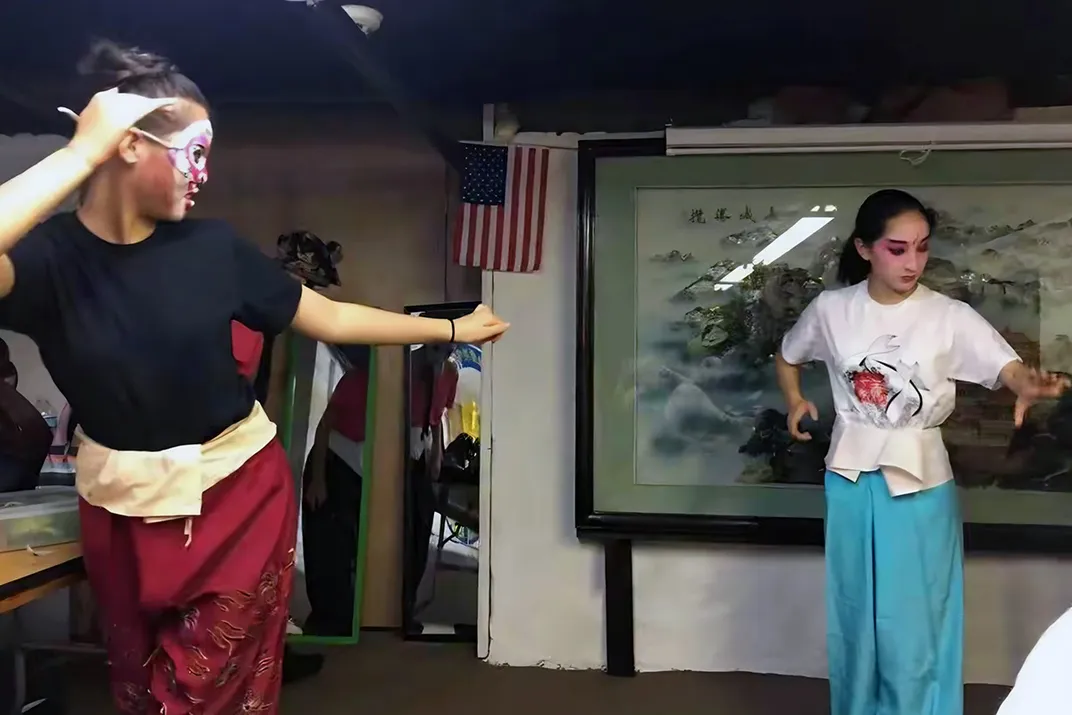 Two teens practice a dance in a dimly lit room, wearing stage makeup and silk pants.