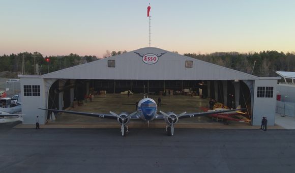 A propellor plane is in front of an open hangar