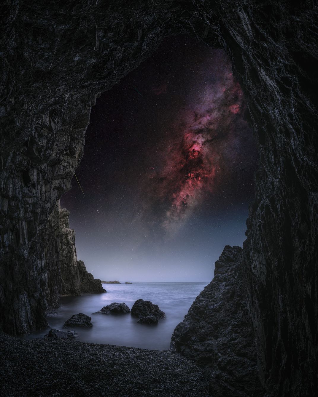 an opening of a cave looks out onto water with a few rounded rocks sticking out, and the sky is lit by a glowing red, starry core of the Milky Way
