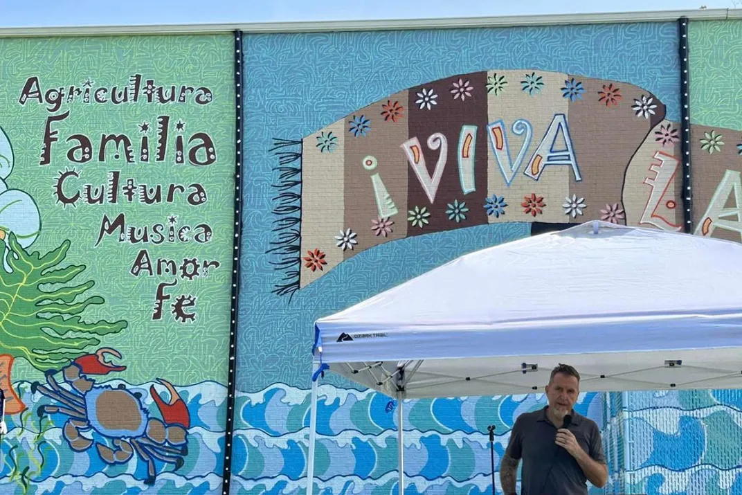 A man speaks into a mic under a white tent. Behind him, the left side of the mural includes words in Spanish: Agricultura, Familia, Cultura, Musica, Amore, along with a blue crab.