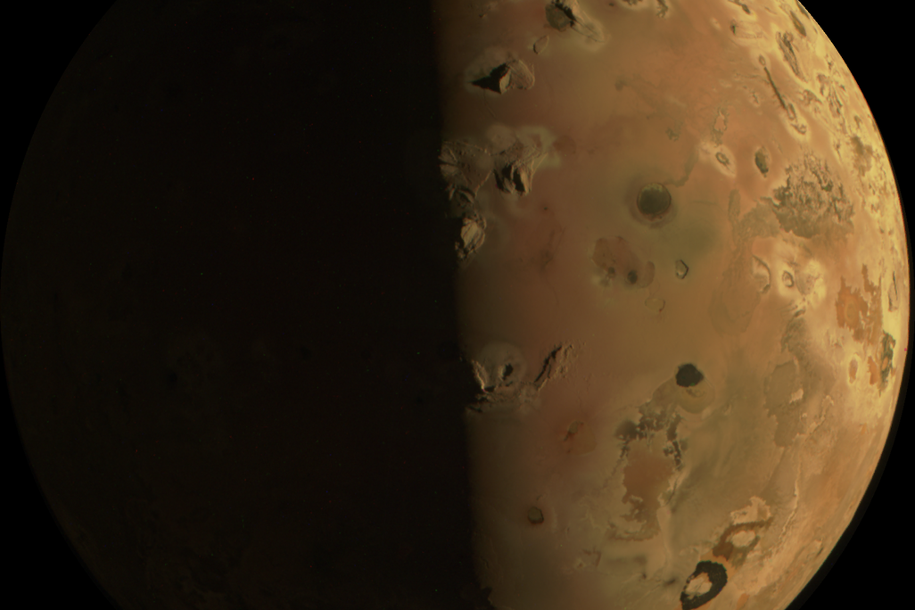 An image of Io, Jupiter's third-largest moon, appearing rust-colored with many visible black pockmarks, which are mountain ranges and surface volcanoes as seen from afar.