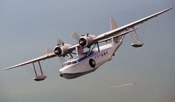 Silver twin engine flying boat over a vast ocean. A small boat leaves it's wake below.