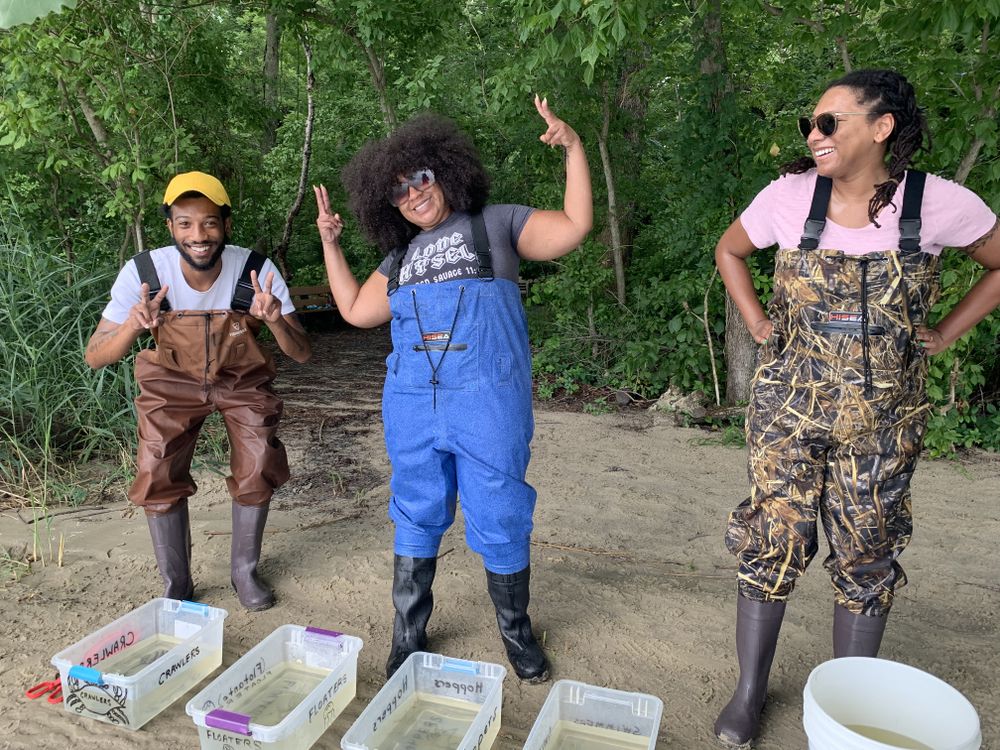 Three adults wearing chest waders laughing and in silly poses. They are standing on sand behind a line of plastic bins filled with water.